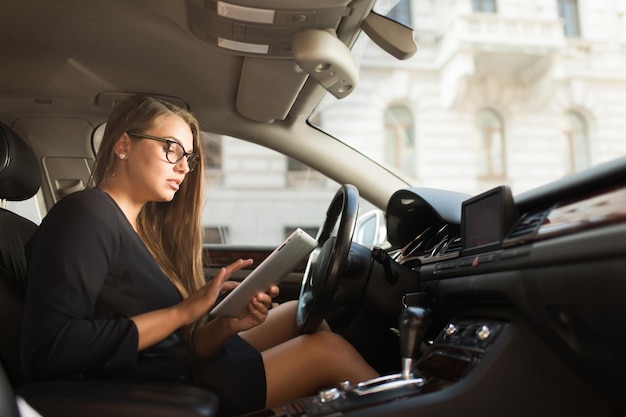 Joven empresaria atractiva con vestido negro y anteojos usando cuidadosamente la tableta mientras se sienta detrás del volante en el auto