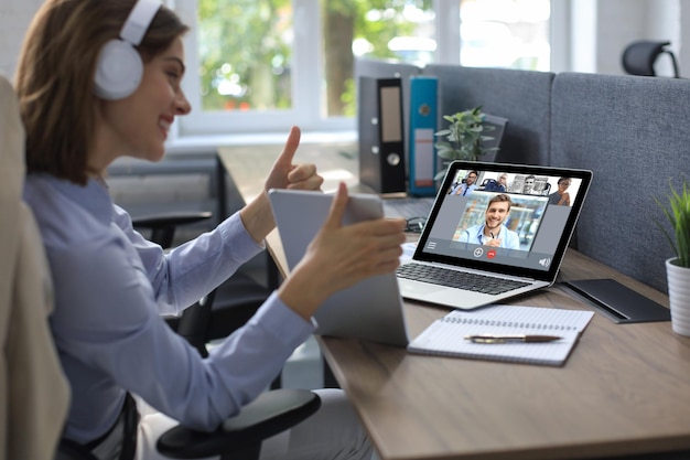 Joven empresaria en auriculares trabajando en PC, haciendo videoconferencia con su colega en la computadora portátil en la oficina. Trabajo a distancia.