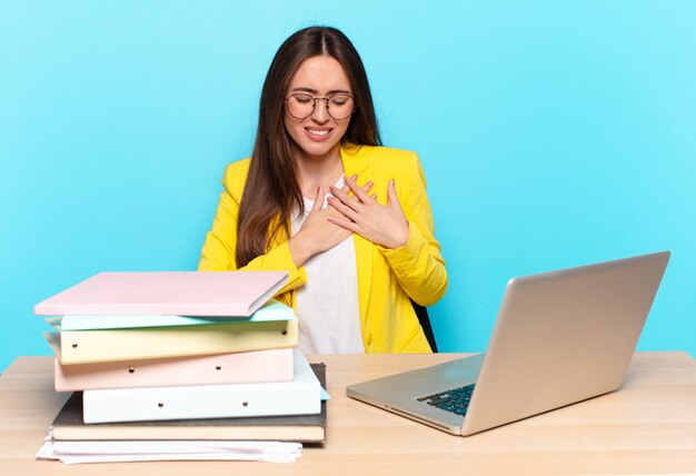 Joven empresaria bonita que parece triste, herida y con el corazón roto, sosteniendo ambas manos cerca del corazón, llorando y sintiéndose deprimida