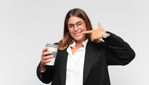 Joven empresaria con un café sonriendo con confianza apuntando a su propia sonrisa amplia, actitud positiva, relajada y satisfecha