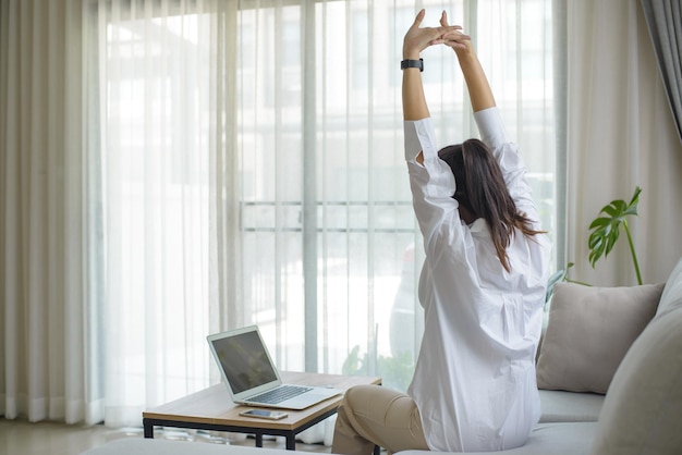 Joven empresaria con camisa blanca sentada en un sofá con una laptop levantando los brazos por encima de la cabeza estirándose para aliviar el dolor después de estar sentada durante mucho tiempo en el trabajo