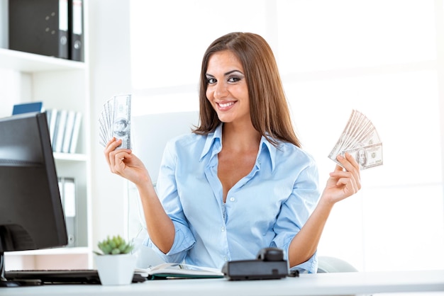 Foto joven empresaria en el cargo, sentada en el escritorio de la oficina, sosteniendo dinero y con una expresión de felicidad en su rostro mirando a la cámara.