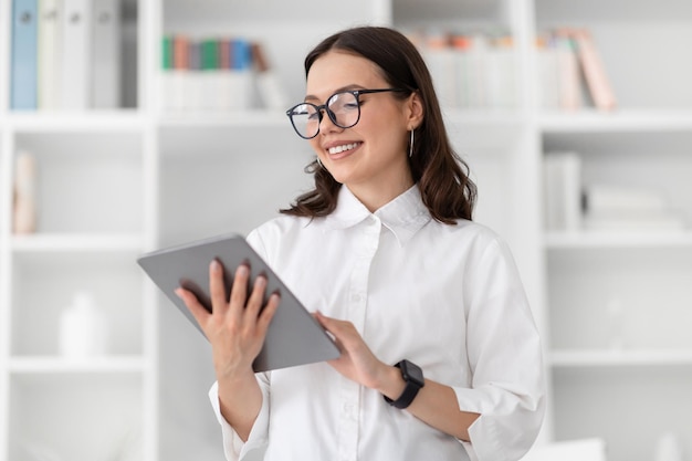 Una joven empresaria caucásica alegre con camisa blanca y gafas charlando en una tableta en el interior de la oficina