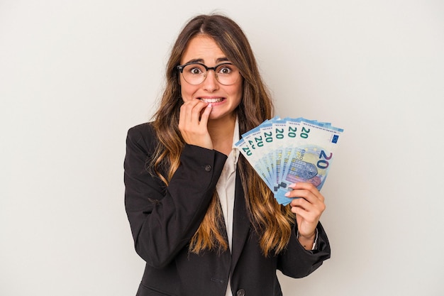 Joven empresaria caucásica sosteniendo billetes aislados de fondo blanco mordiéndose las uñas, nerviosa y muy ansiosa.