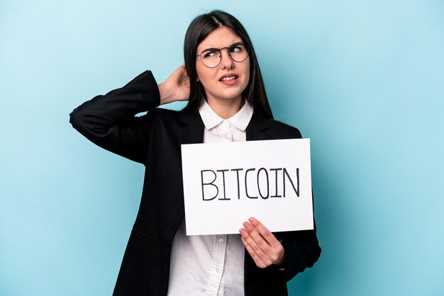 Joven empresaria caucásica sosteniendo un cartel de bitcoin aislado de fondo azul tocando la parte posterior de la cabeza pensando y tomando una decisión