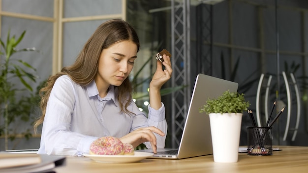 Joven empresaria comiendo un donut glaseado mientras trabaja en una laptop en la oficina