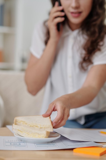 Joven empresaria comiendo sándwich cuando habla por teléfono con un colega y comenta el último informe