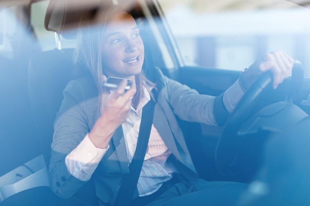 Joven empresaria conduciendo un auto y comunicándose por teléfono celular La vista es a través del vidrio