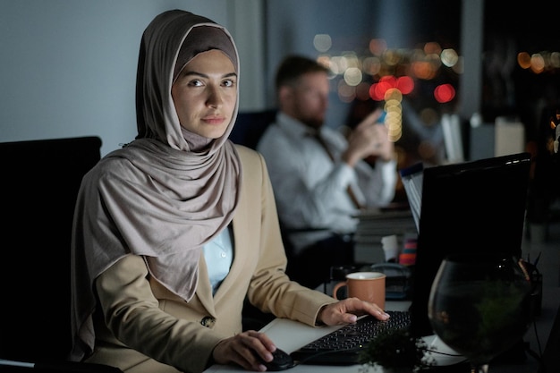 Joven empresaria contemporánea en hijab mirando a la cámara mientras está sentado frente a la computadora