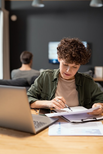 Joven empresaria contemporánea mirando a través del texto del contrato mientras está sentada junto a la mesa frente a la computadora portátil contra su esposo tranquilo