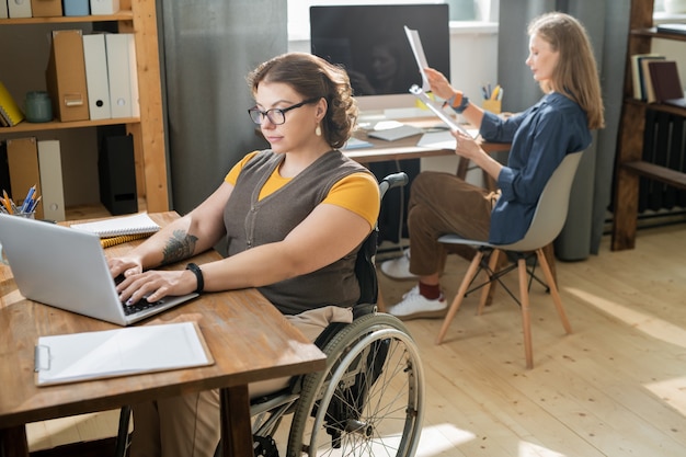 Foto joven empresaria discapacitada sentada en silla de ruedas frente a la computadora portátil y desplazándose a través de los datos en línea sobre los antecedentes de su ocupado colega