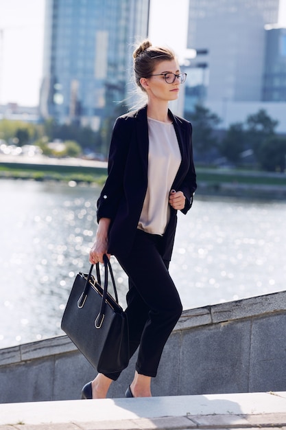 Joven empresaria elegante con bolso de cuero negro moviéndose arriba frente a la cámara contra la orilla del río y un grupo de edificios modernos