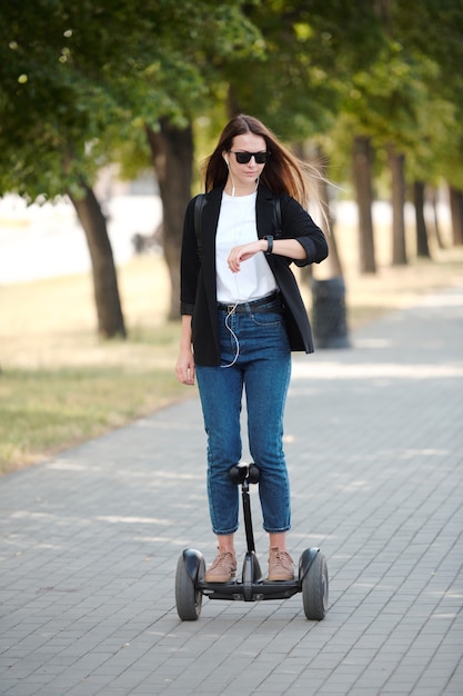 Joven empresaria elegante contemporánea mirando el reloj de pulsera mientras viaja en giroscopio a lo largo de la carretera contra árboles verdes que crecen en fila