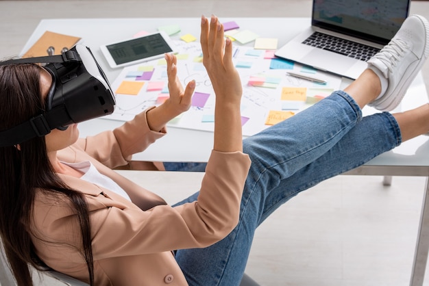 Joven empresaria elegante en ropa casual elegante y auriculares vr sentada junto al escritorio y preparando la presentación mientras toca la pantalla virtual