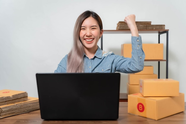 Una joven empresaria está muy feliz después de revisar su correo electrónico a través de su computadora portátil. Los vendedores en línea están empaquetando productos en cajas de paquetes.