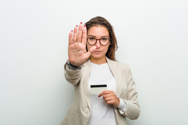 Joven empresaria europea sosteniendo una tarjeta de crédito de pie con la mano extendida mostrando la señal de stop, impidiéndole.