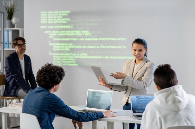 Joven empresaria exitosa apuntando a la pantalla del portátil mientras explica datos a sus colegas durante la presentación de su proyecto