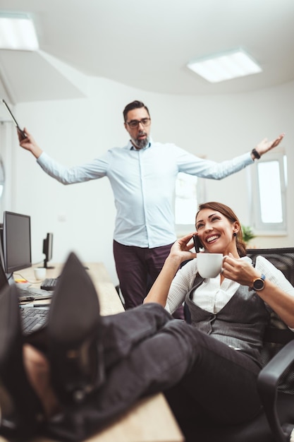 Una joven empresaria exitosa está hablando por un teléfono inteligente en la oficina y su colega está impaciente por continuar trabajando.