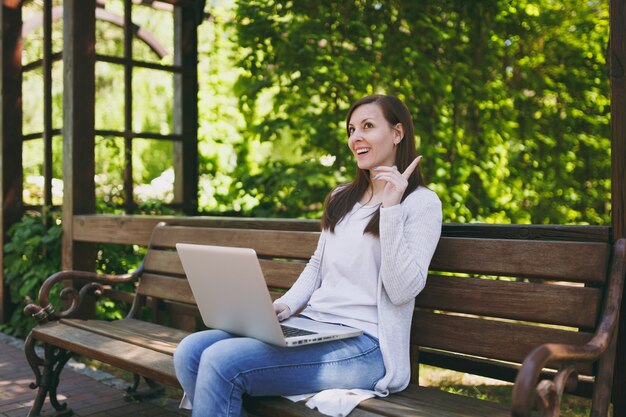 Joven empresaria exitosa en ropa casual ligera. Mujer sentada en un banco trabajando en un moderno ordenador portátil en el parque de la ciudad en la calle al aire libre en la naturaleza. Oficina móvil. Concepto de negocio autónomo