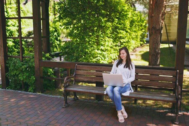 Joven empresaria exitosa en ropa casual ligera. Mujer sentada en un banco trabajando en un moderno ordenador portátil en el parque de la ciudad en la calle al aire libre en la naturaleza. Oficina móvil. Concepto de negocio autónomo