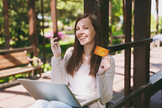 Joven empresaria exitosa con tarjeta de crédito. Mujer sentada en un banco trabajando en un moderno ordenador portátil en el parque de la ciudad en la calle al aire libre en la naturaleza. Oficina móvil. Freelance, concepto de negocio.