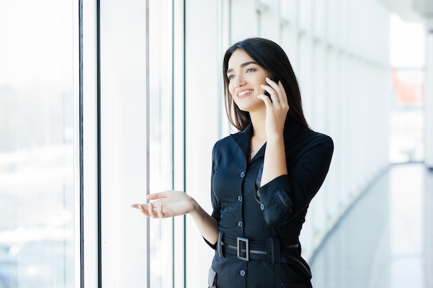 Joven empresaria hablando por teléfono móvil mientras está de pie junto a la ventana en la oficina. Modelo de mujer joven hermosa en oficina brillante.