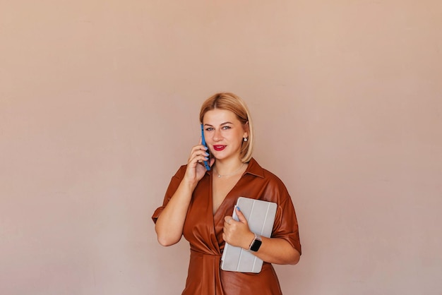 Joven empresaria hermosa con un vestido marrón hablando por teléfono con una computadora portátil en sus manos Joven empresaria alegre está teniendo una conversación agradable