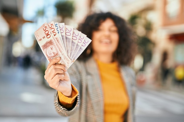 Joven empresaria hispana sonriendo feliz sosteniendo billetes de coronas suecas en la ciudad