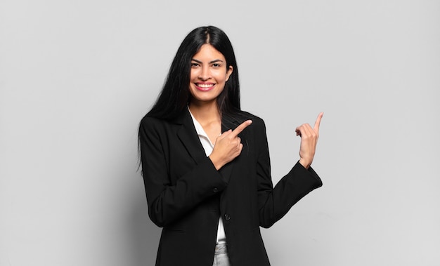 Joven empresaria hispana sonriendo felizmente y apuntando hacia un lado y hacia arriba con ambas manos mostrando el objeto en el espacio de la copia