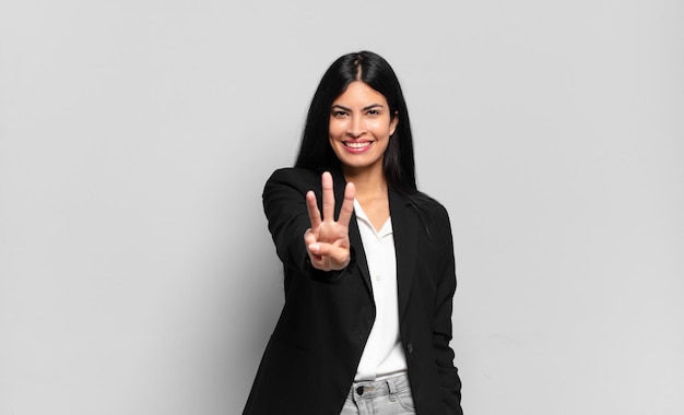 Joven empresaria hispana sonriendo y mirando amistosamente, mostrando el número tres o tercero con la mano hacia adelante, contando hacia atrás