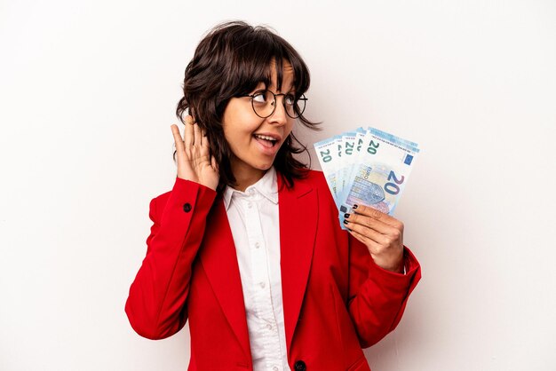 Joven empresaria hispana sosteniendo billetes aislados de fondo blanco tratando de escuchar un chisme