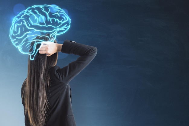 Joven empresaria con holograma de cerebro digital brillante de pie sobre fondo de pared de pizarra borrosa con lugar de maqueta El concepto de proceso de pensamiento de trabajo de cerebro de inteligencia