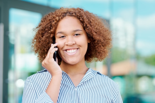 Joven empresaria intercultural alegre con cabello castaño rizado hablando por teléfono inteligente después del trabajo en entorno urbano