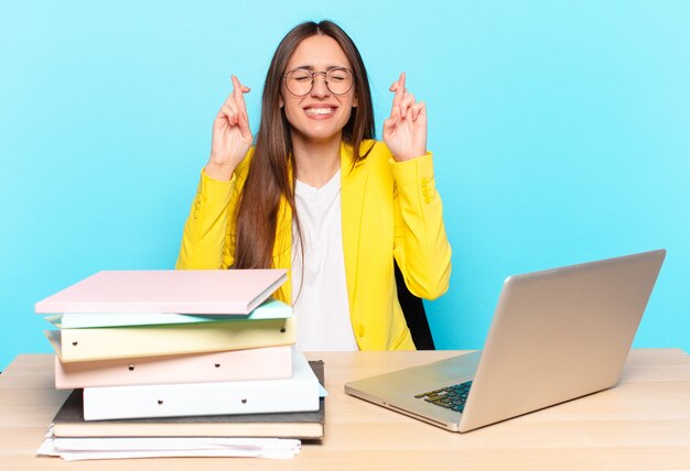 Joven empresaria joven sonriendo y cruzando ansiosamente ambos dedos, sintiéndose preocupada y deseando o esperando buena suerte