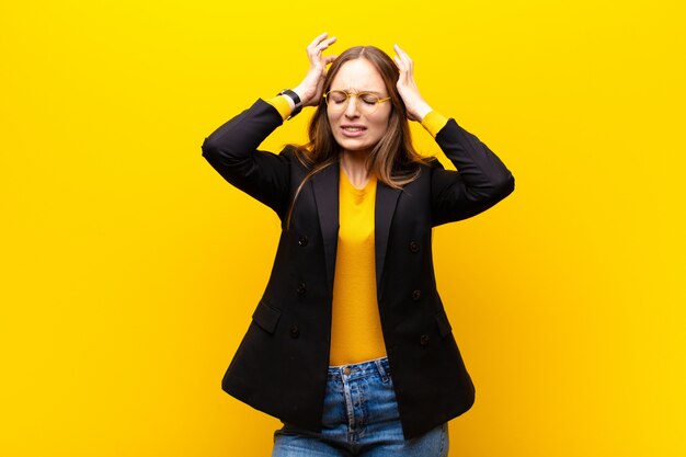 Joven empresaria linda sintiéndose estresada y frustrada, levantando las manos a la cabeza, sintiéndose cansada, infeliz y con migraña contra la pared naranja