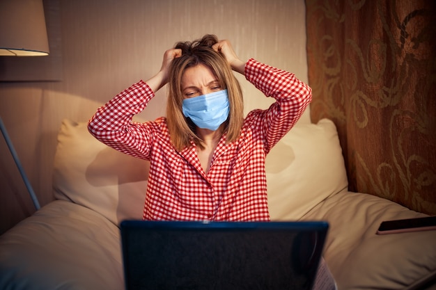 Foto joven empresaria en una máscara protectora médica trabaja desde su casa en la computadora durante el autoaislamiento y la cuarentena.