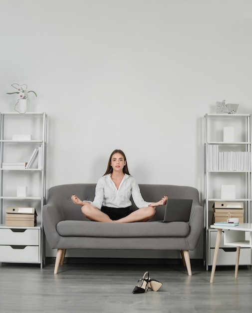 Joven empresaria de meditación haciendo ejercicio de yoga en el sofá en el lugar de trabajo en un empleado de oficina moderno