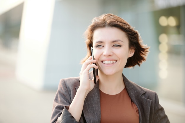 Joven empresaria morena sonriente en cliente de consultoría de ropa casual inteligente en el teléfono contra el exterior de la arquitectura contemporánea