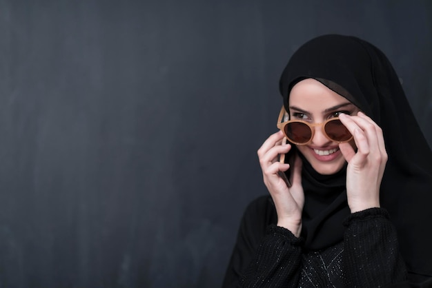 Joven empresaria musulmana moderna que usa smartphones con gafas de sol y ropa hijab frente a una pizarra negra.