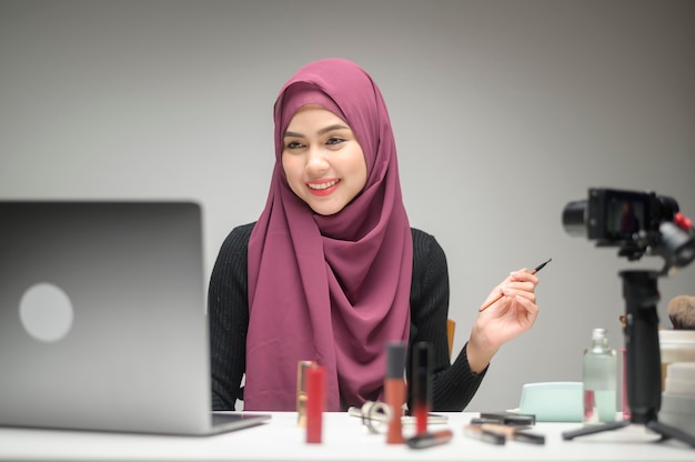 Una joven empresaria musulmana que trabaja con una computadora portátil presenta productos cosméticos durante la transmisión en vivo en línea sobre un estudio de fondo blanco, vendiendo en línea y concepto de blogger de belleza