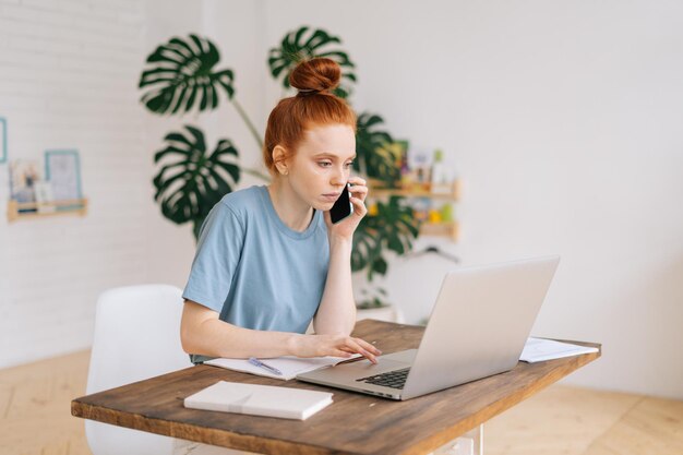 Una joven empresaria pelirroja seria que trabaja en una computadora portátil en el escritorio y habla por teléfono móvil en una habitación luminosa y acogedora en la oficina del hogar. Concepto de trabajo a distancia en Internet en autoaislamiento.