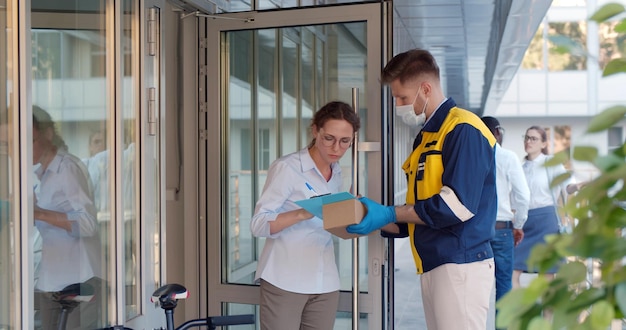 Joven empresaria que firma el documento en el portapapeles recibe el paquete del servicio de mensajería con máscara de seguridad