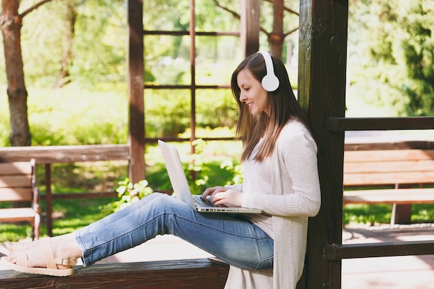 Joven empresaria en ropa casual ligera. Mujer que trabaja en la computadora portátil moderna, escuchar música con auriculares en la cabeza en la calle al aire libre. Oficina móvil. Concepto de negocio autónomo. Vista lateral.