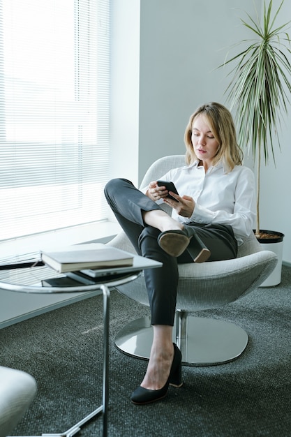 Joven empresaria rubia elegante con gadget móvil sentado en un cómodo sillón junto a la ventana y una mesa pequeña con un cuaderno