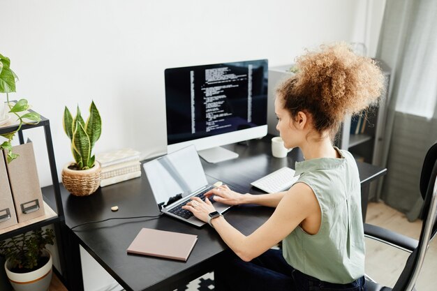 Joven empresaria sentada en la mesa frente al monitor de la computadora y escribiendo en la computadora portátil en la oficina