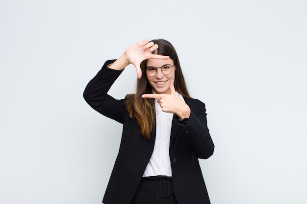 Joven empresaria se siente feliz, amigable y positiva, sonriendo y haciendo un retrato o marco de fotos con las manos en la pared blanca