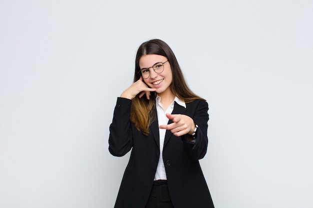 Joven empresaria sonriendo alegremente y señalando mientras hace una llamada más tarde gesto, hablando por teléfono contra la pared blanca