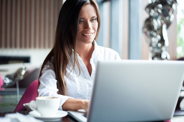 Foto joven empresaria sonriente en un descanso en un café. ella está trabajando en una computadora portátil y tomando café.