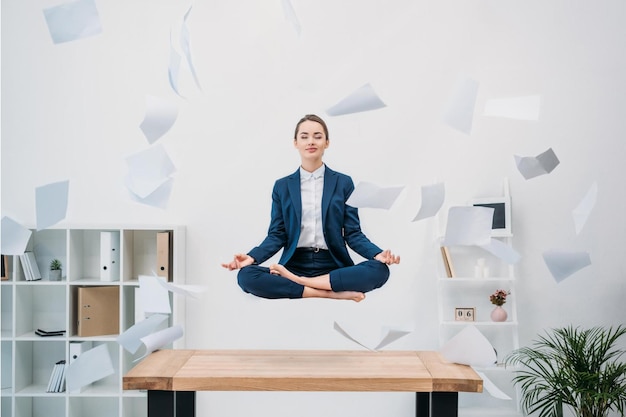 Joven empresaria sonriente con los ojos cerrados meditando mientras levita en el lugar de trabajo con papeles