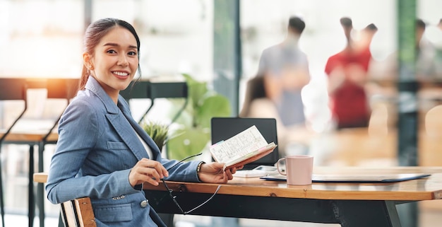 Una joven empresaria sonriente con traje que sostiene una nota de plan de negocios y se sienta en el escritorio en una moderna oficina de coworking